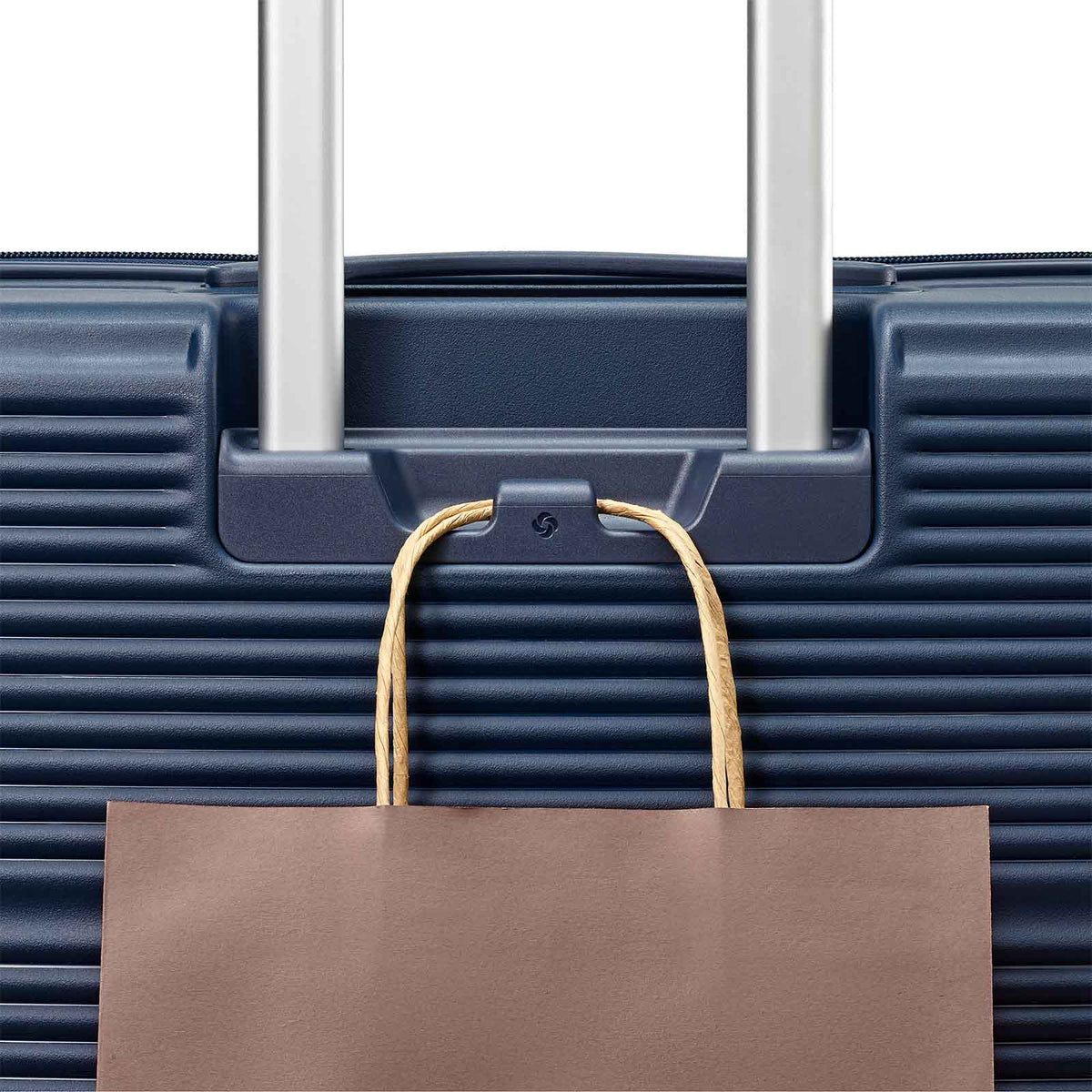 Close-up of a dark blue hard-shell suitcase with silver telescoping handles and a brown paper shopping bag hanging from a hook on the front.