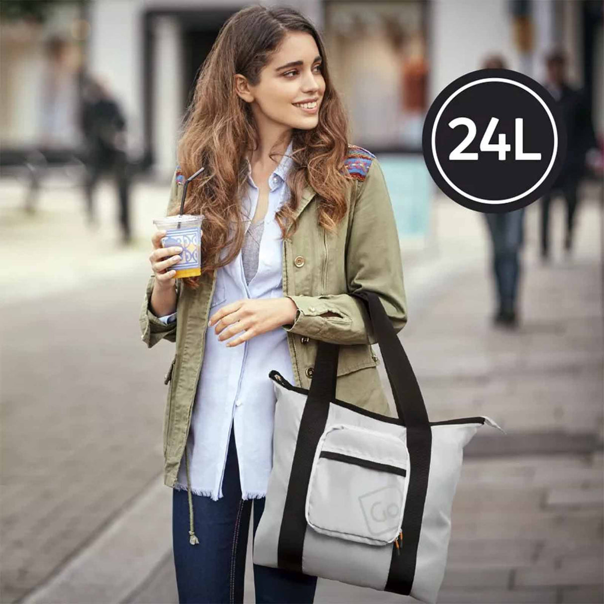 Woman holding a white bag with black straps and a drink, standing on a street.