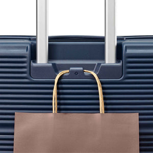 Close-up of a dark blue hard-shell suitcase with silver telescoping handles and a brown paper shopping bag hanging from a hook on the front.