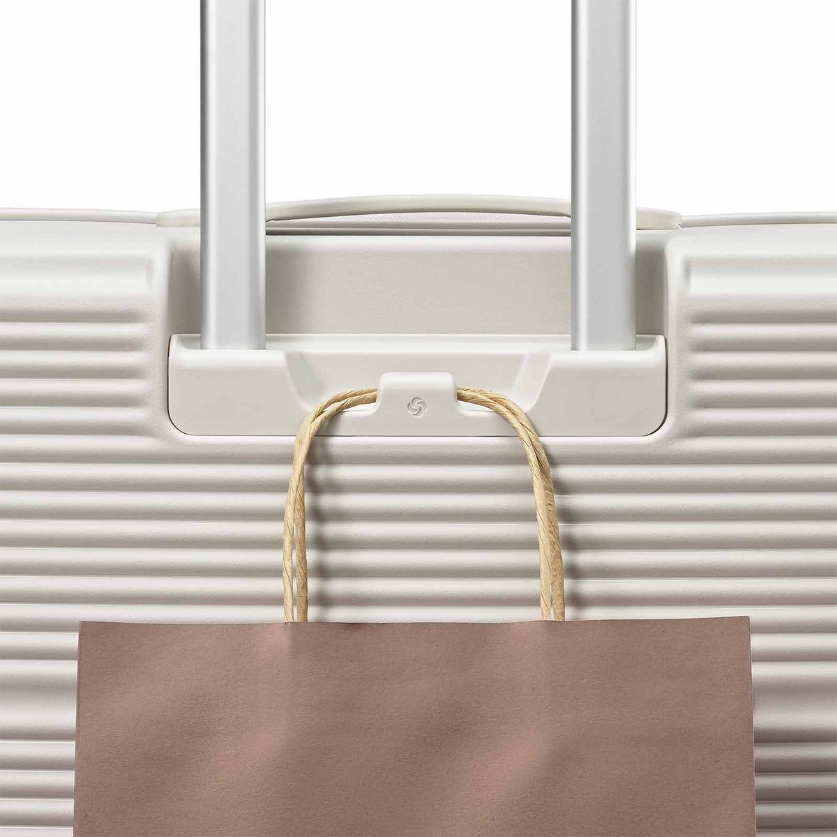 A close-up of a white hard-shell suitcase with extended silver handles and a brown paper shopping bag hanging from a hook on the front.