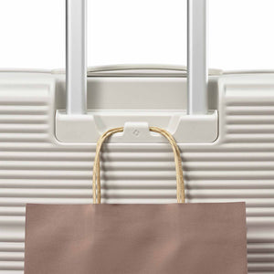 A close-up of a white hard-shell suitcase with extended silver handles and a brown paper shopping bag hanging from a hook on the front.