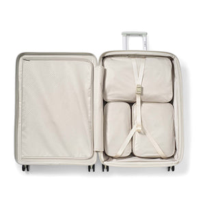 An open hard-shell suitcase in a light beige or off-white color, shown from a top-down view. Inside, four matching beige packing cubes are arranged and secured with a white bungee strap.