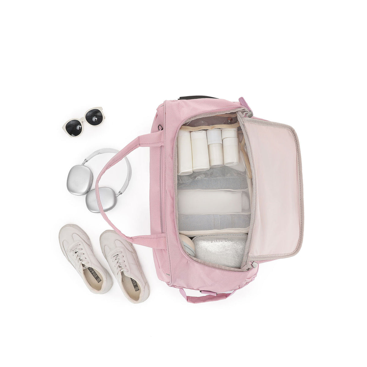 A pink Carry-On bag shown from above with its interior open, filled with travel items including white toiletry bottles, a gray organizer insert, and a small cooler. White sneakers, silver headphones, and black sunglasses are placed beside it on a clean white background.