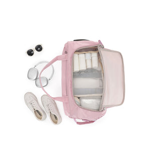 A pink Carry-On bag shown from above with its interior open, filled with travel items including white toiletry bottles, a gray organizer insert, and a small cooler. White sneakers, silver headphones, and black sunglasses are placed beside it on a clean white background.